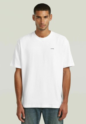 Homme aux cheveux courts et foncés portant un t-shirt blanc uni oversize et un jean bleu sur un fond clair.