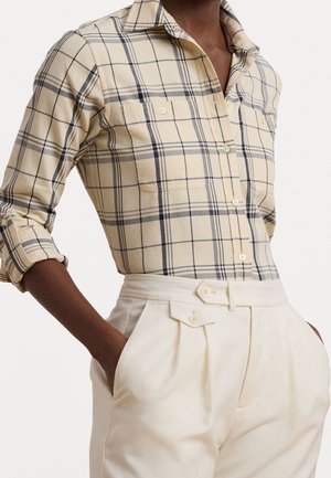 Persoon draagt een beige geruite blouse met opgerolde mouwen, ingestopt in een hoge taille crèmebroek met knoopdetails.