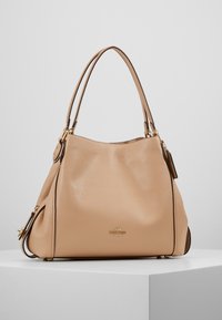 Sac seau en cuir beige avec deux poignées, surface texturée et accents en métal doré. Présente une petite étiquette de marque.