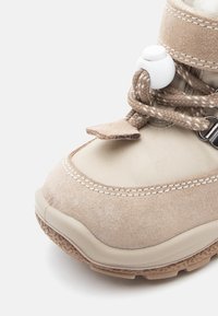 Botină bej impermeabilă cu partea superioară din suede și material sintetic, talpă din cauciuc întărită, șireturi elastice și închidere prin toggle pentru o fixare sigură.