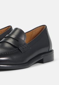 Mocassins en cuir noir avec une finition lisse, affichant une sangle décorative et un faible talon. À l'intérieur, une doublure beige pour plus de confort.