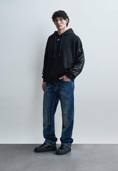 Jauns vīrietis stāv pret baltu sienu, valkājot melnu kapuci ar baltu tekstu un zilas džinsas ar aizšūtām vietām, kā arī melnas sporta kurpes.