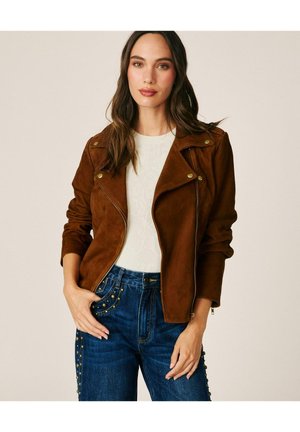 Mujer con cabello largo y oscuro que lleva una chaqueta de ante marrón sobre una camiseta blanca y vaqueros azules con tachuelas de latón en los bolsillos y los laterales.