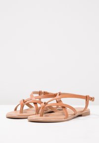 Tan strappy sandals with a flat sole, featuring crisscross straps and a buckled ankle strap. Smooth texture and minimalist design.