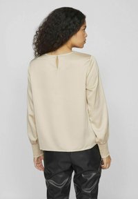 Beige långärmad blus med detalj i nyckelhål bak och texturerade smockade manschetter. Kombinerad med svarta läderbyxor som visar en jämn tyg.