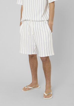 Homme portant un short blanc et une chemise assortie à fines rayures bleues, les mains dans les poches, et des tongs blanches sur fond gris.