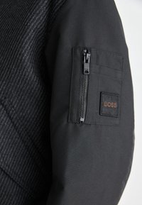 Veste noire avec un tissu rayé texturé d'un côté et un matériau lisse de l'autre. Elle possède une poche zippée et un écusson de logo.