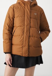 Brown puffer jacket with a hood, featuring quilted sections, zip closure, and logo detail. Worn over a black skirt. Soft texture.