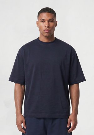 Homme aux cheveux courts portant un t-shirt uni bleu foncé oversize et un pantalon assorti, debout devant un fond clair.