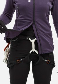Personne portant un harnais d'escalade noir avec des boucles orange et une veste violette, ajustant la fermeture éclair de la veste sur un fond blanc.