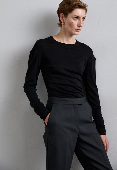 Woman with short hair wearing gold hoop earrings, a black long-sleeve top, and high-waisted dark gray trousers, standing against a gray wall.