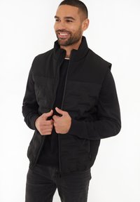Schwarze Steppweste mit hohem Kragen, seitlichen Reißverschlusstaschen und gestepptem Design, getragen über einem schwarzen Sweatshirt. Das Model steht lächelnd.
