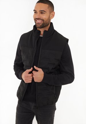 Zwarte puffer vest met hoge kraag, zijrits zakken en gewatteerde textuur, gedragen over een zwarte sweatshirt. Model staat glimlachend.