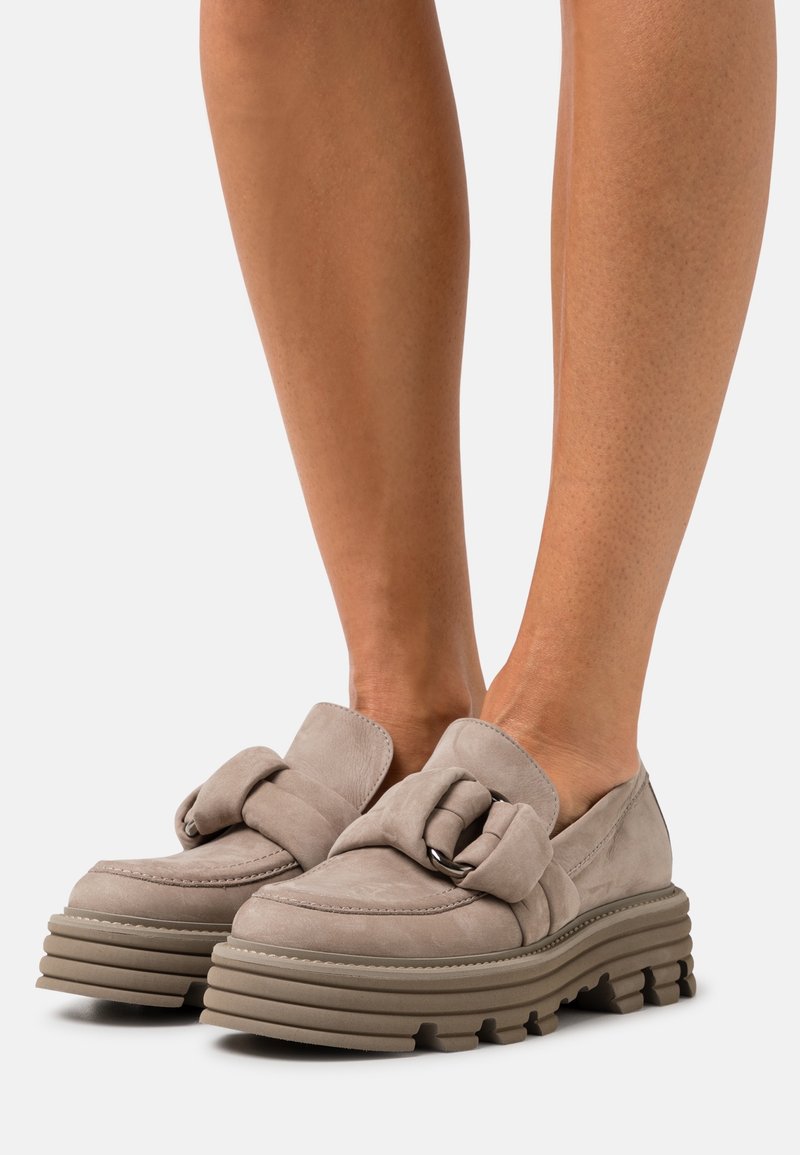 Mocassins beiges en daim épais avec semelles crantées épaisses et grand détail de bride nouée, portés pieds nus.