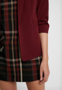 Burgundy blazer with a smooth texture and rolled sleeves, paired with a plaid mini skirt in black, red, and beige tones.