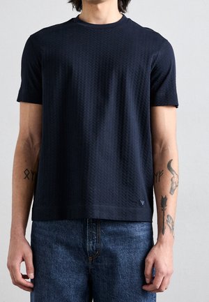Torso d'homme mince portant un t-shirt noir à manches courtes texturé et un jean bleu, montrant des tatouages sur les avant-bras contre un fond uni.