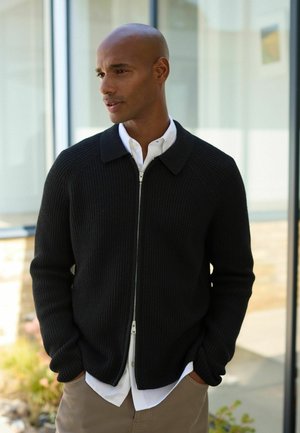 Man with shaved head wearing a black zip-up sweater over a white shirt and beige pants standing indoors near a glass window.