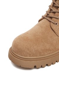 Hnědé semišové boty s kulatou špičkou a texturovaným povrchem. Obsahují silnou gumovou podrážku s horizontálními rýhami a kovovými očky na tkaničky.