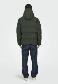 Chaqueta de plumas en verde oscuro con capucha, que presenta secciones acolchadas y un corte recto, combinada con jeans de mezclilla azules y zapatillas blancas.