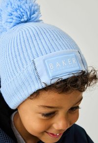 Svijetloplava pletena kapa s pom-pomom na vrhu i pravokutnom etiketom na kojoj piše "BAKER by TED BAKER" ušivenom bijelom bojom.