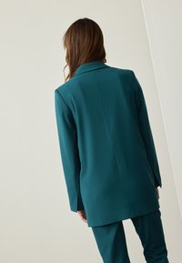 Teal blazer with a tailored fit, notched lapels, and a smooth texture, paired with matching trousers. Features a straight back design.