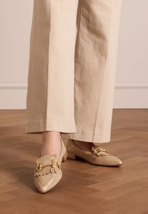 Beige loafers with a pointed toe, featuring fringe detail and gold hardware accents. Low heel and suede texture, displayed on a wooden floor.