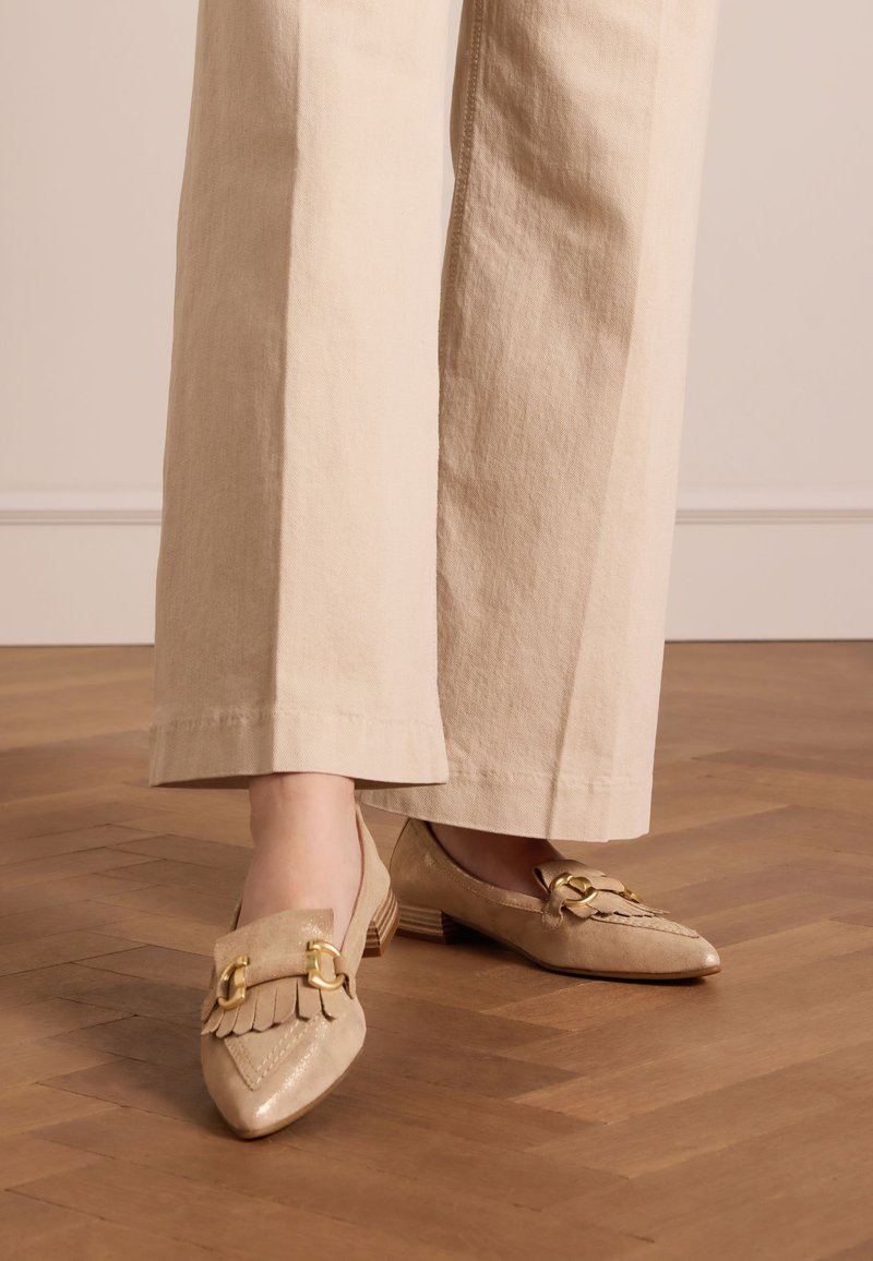 Beige Loafer mit spitzem Toe, mit Fransen und Goldbeschlägen. Niedriger Absatz und Wildleder-Optik, auf einem Holzboden präsentiert.