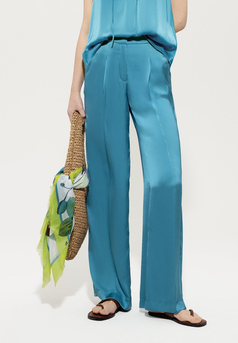 Helle blaue Satin-Hose mit lässiger Passform, flachem Bund und seitlichen Taschen, kombiniert mit einer gewebten Strohtasche und einem grünen Blumen-Schal.