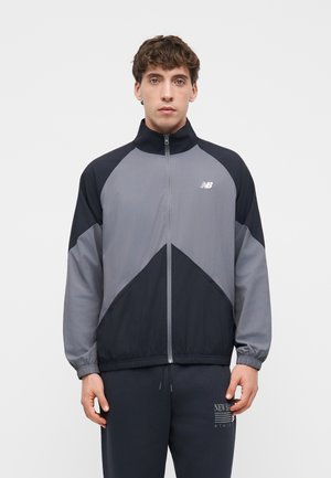Ung mand iført en grå og sort lynlåslukket New Balance træningsjakke og sorte joggingbukser mod en ensfarvet grå baggrund.