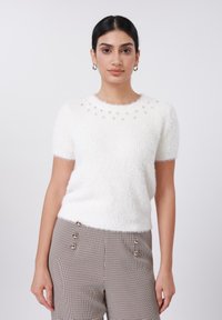 Pull blanc à manches courtes et pelucheux avec des ornements autour de l'encolure. Porté avec un pantalon à carreaux orné de boutons décoratifs.