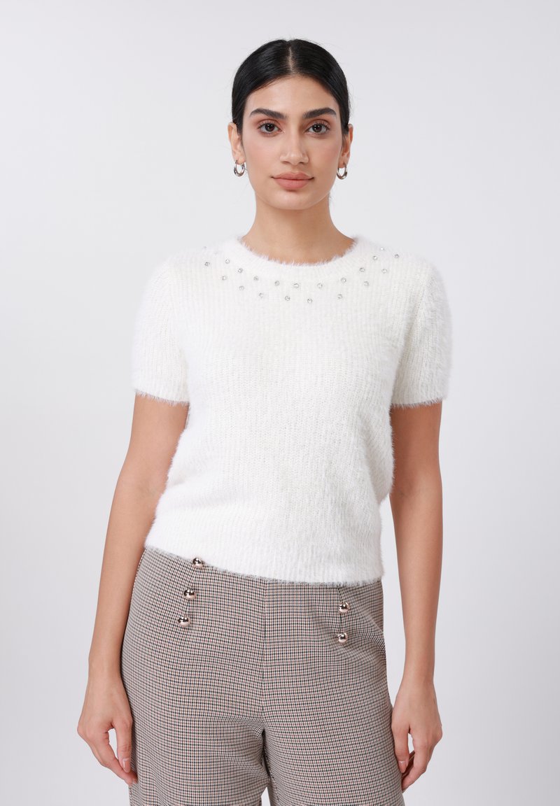 Pull blanc à manches courtes et pelucheux avec des ornements autour de l'encolure. Porté avec un pantalon à carreaux orné de boutons décoratifs.