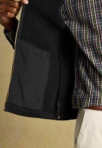 Melnā flīša oderejā jaka ar rāvējslēdzēju, aprīkota ar iekšējām kabatām, regulējamu apakšmalu un gludu ārējo virsmu.