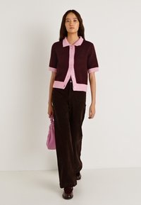 Καρδινάλια και ροζ κοντομάνικο ζακέτα με κουμπιά, συνδυασμένη με σκούρο καφέ παντελόνια φαρδιάς γραμμής. Το μοντέλο κρατά ροζ τσάντα χειρός.