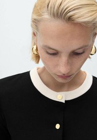 Maglietta nera con colletto crema, con bottoni dorati. I capelli corti biondi del modello sono visibili. Lo sfondo è semplice e di colore chiaro.