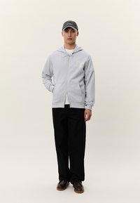 Mladý muž oblečený v svetlosivej mikine na zips s logom, čierne široké nohavice, tmavohnedé topánky a čierna čiapka, stojaci pred jednoduchým pozadím.