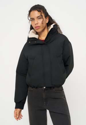 Mujer con cabello rizado que lleva una chaqueta acolchada negra con cuello alto forrado de borreguito beige y vaqueros gris oscuro.