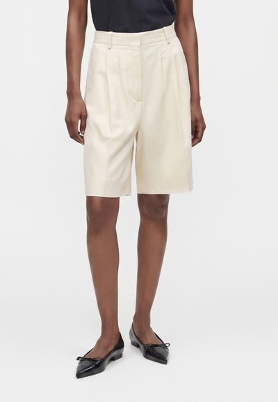 Personne portant un short crème taille haute, longueur genou, taillé sur mesure, associé à des ballerines noires à bout pointu et un haut foncé sur fond uni.