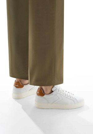 Weiße Leder-Sneaker mit braunem Fersenakzent, runder Spitze, flacher Gummisohle und strukturierten Oberflächen. Kombiniert mit weiten olivfarbenen Hosen.