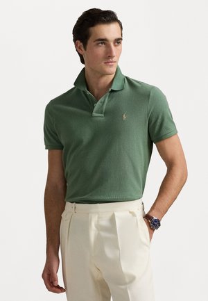 Camisa polo verde, de manga corta, con tejido texturizado, cuello clásico, logotipo bordado en dorado. Combinada con pantalones beige claros a medida.