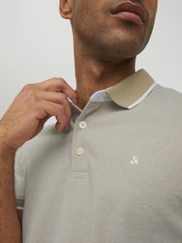 Beige Polohemd aus strukturiertem Stoff, mit einem hellgrauen Kragen und drei Knöpfen. Es hat ein kleines weißes Logo auf der Brust.