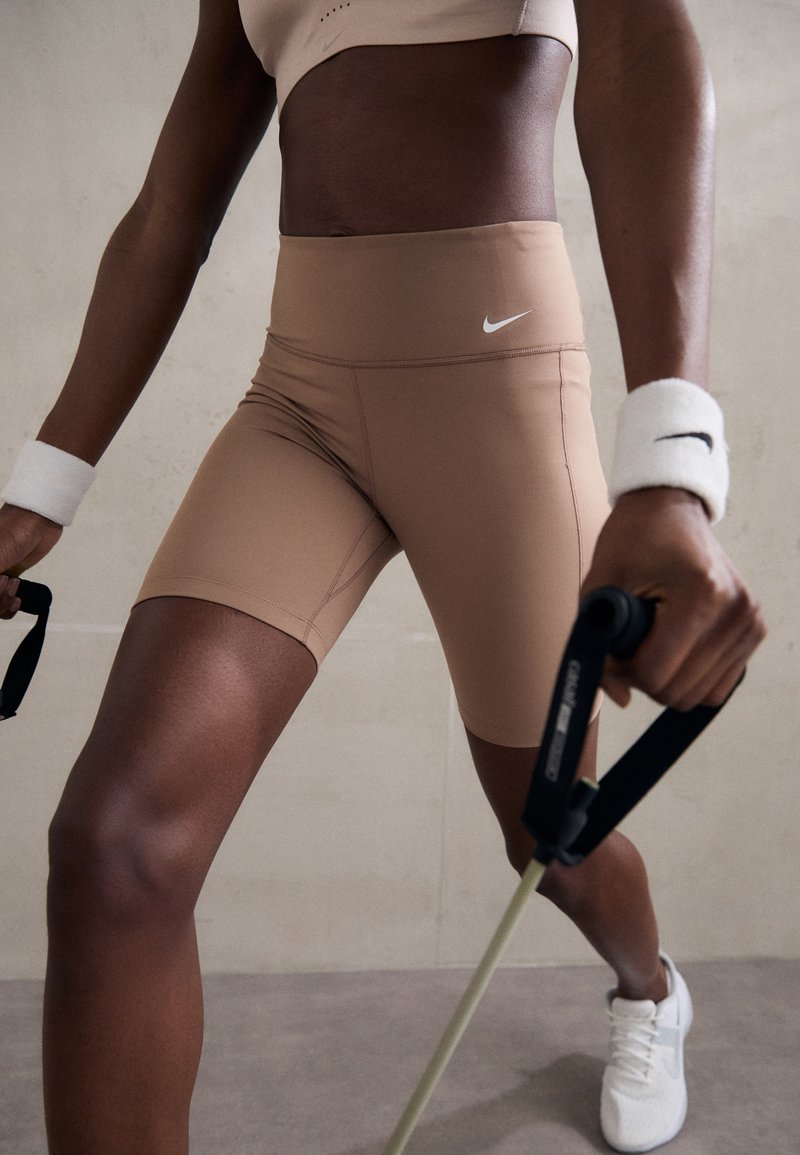 Personne portant un short Nike beige et des bandeaux blancs aux poignets, utilisant des bandes de résistance en position d'entraînement sur un fond neutre.