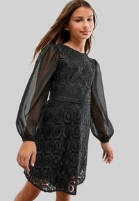 Fille portant une robe noire en dentelle avec des motifs circulaires et des manches longues transparentes à volants, se tenant devant un fond gris uni.
