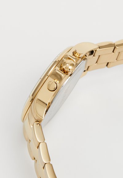 Montre en métal doré avec un boîtier lisse et arrondi, un bracelet texturé et des boutons de pression sur le côté. Le fond est argenté avec des détails gravés.