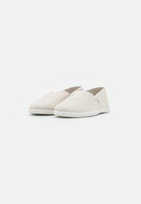Sapatilhas slip-on de lona bege clara com parte superior texturizada, detalhes elásticos e solas de borracha brancas. Design minimalista, sem padrões visíveis.