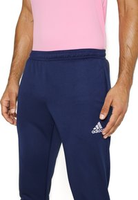 Pantaloni sportivi blu navy con fascia elastica in vita, caratterizzati da un logo bianco Adidas sulla parte bassa della gamba sinistra. Realizzati in tessuto liscio e traspirante.