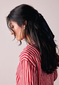 Rot-weiß gestreiftes Hemd mit Kragen, getragen von einer Person mit langen schwarzen Haaren, die mit einem marineblauen Band gebunden sind. Dezenter Ohrring sichtbar.