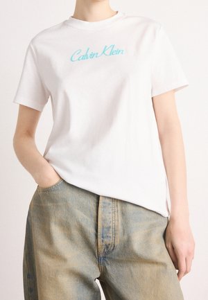 Bílé tričko s krátkým rukávem s světle modrým nápisem "Calvin Klein", nošené s vybledlými modrými džínami s vysokým pasem, jedna ruka v kapse.