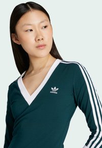 Zelený dlhý rukáv s výstrihom do V, bielym golierom a logom Adidas. Obsahuje biele pruhy pozdĺž rukávov, vyrobené z mäkkého materiálu.