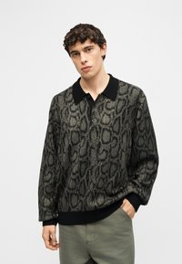 Νέος άνδρας με σγουρά μαλλιά που φοράει γκρι και μαύρο πουκάμισο πόλο με μοτίβο φιδιού και ανοιχτό γκρι παντελόνι, με το χέρι στην τσέπη.