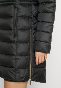 Schwarzer Steppmantel mit gesteppter Textur, einer Vordertasche mit Reißverschluss und einem goldfarbenen Reißverschlussdetail an der Seite. Isoliert für Wärme.